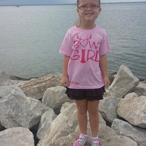 May include: A young girl wearing a pink t-shirt with the text "Bow Girl" in red lettering and a heart. She is standing on rocks by the water wearing pink and white sneakers.