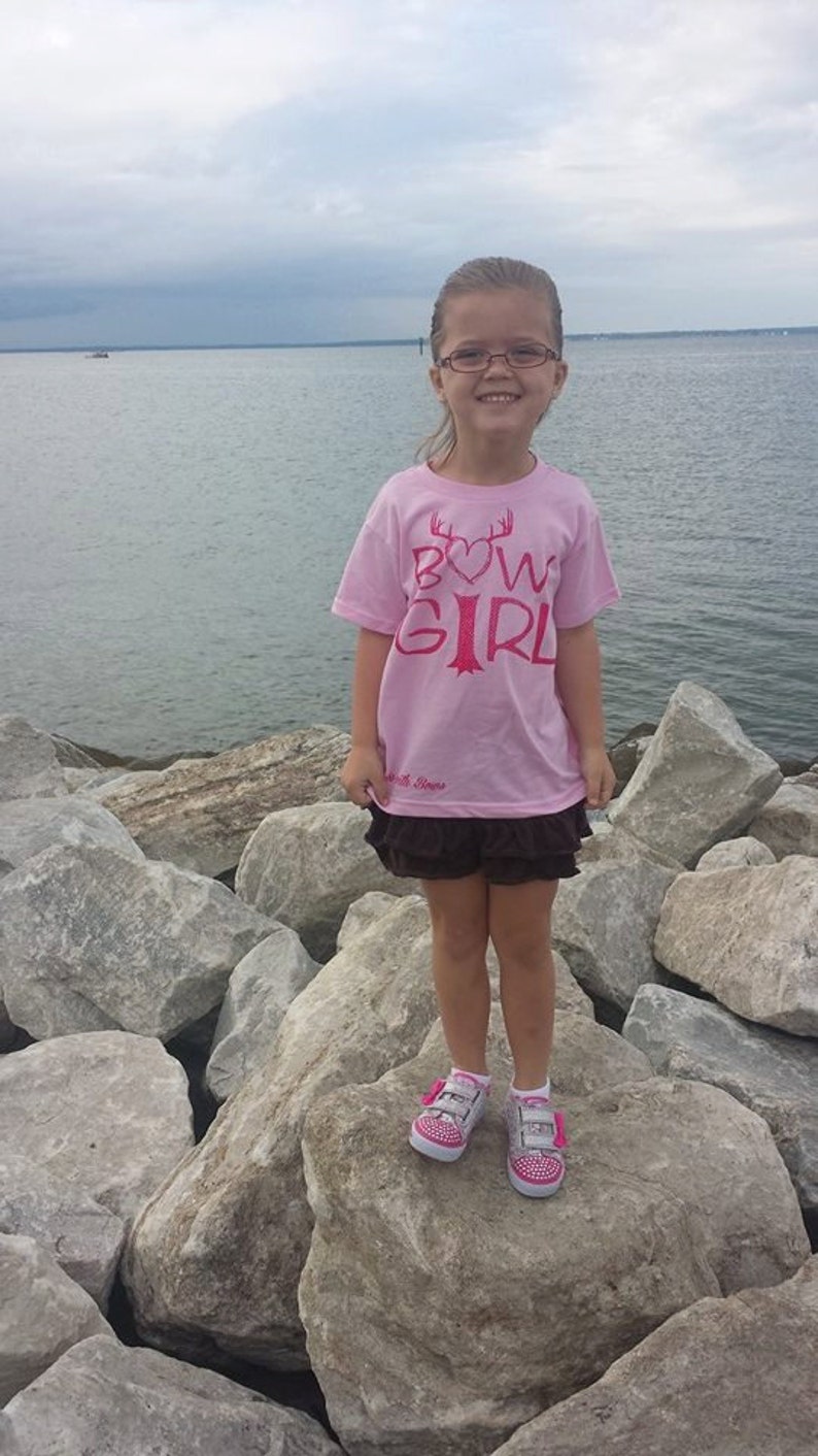 May include: A young girl wearing a pink t-shirt with the text "Bow Girl" in red lettering and a heart. She is standing on rocks by the water wearing pink and white sneakers.