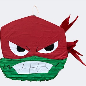 May include: Red and green Teenage Mutant Ninja Turtles piñata with a fierce expression. The piñata is shaped like a turtle head with a red bandana and a green mask.