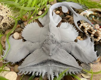 May include: A gray 3D printed model of a fish with a large mouth and a frilly head. The fish is posed with its mouth open and its fins spread out. The model is sitting on a surface with some rocks and plants.