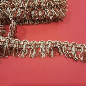 May include: Decorative fringe trim in shades of brown and cream. The trim features a looped design with hanging tassels. A roll of the same trim is visible in the background. This trim is suitable for various craft and sewing projects.