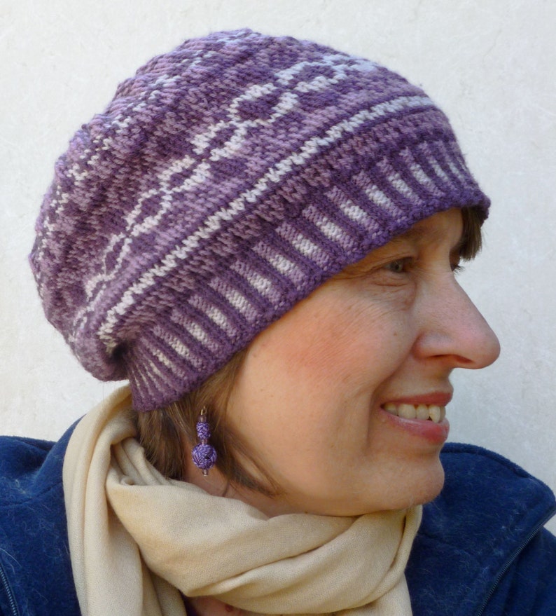 May include: A purple and white knitted beanie with a geometric pattern. The beanie is worn by a person with a light brown scarf and a blue jacket.