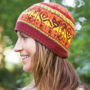 May include: A knitted beanie hat with a colorful geometric pattern. The hat features a repeating design in shades of orange, yellow, and brown, with a red and green striped brim. The hat is worn by a person with long, light brown hair.