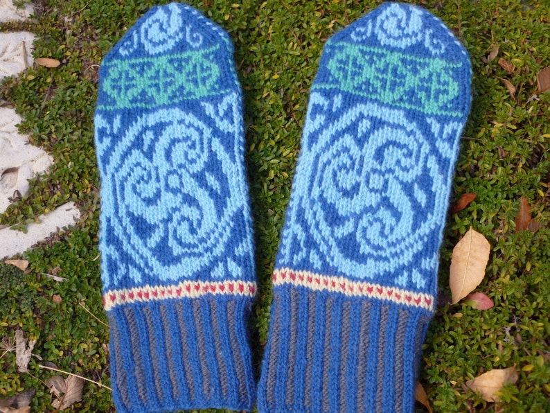 May include: A pair of blue knitted mittens with a white and green patterned design. The mittens have a ribbed cuff and a decorative band of red, white, and cream.
