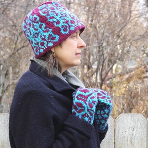 May include: A woman wearing a dark blue coat with a burgundy and teal patterned knit hat and matching mittens.