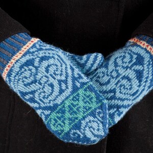 May include: A pair of blue, green, and white knitted mittens with a geometric pattern. The mittens are folded over each other and are being held by a person wearing a black coat.