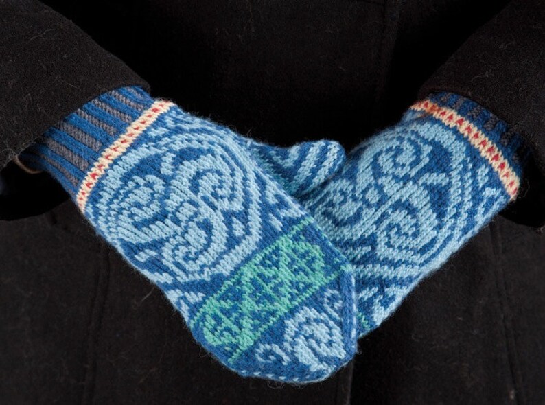 May include: A pair of blue, green, and white knitted mittens with a geometric pattern. The mittens are folded over each other and are being held by a person wearing a black coat.