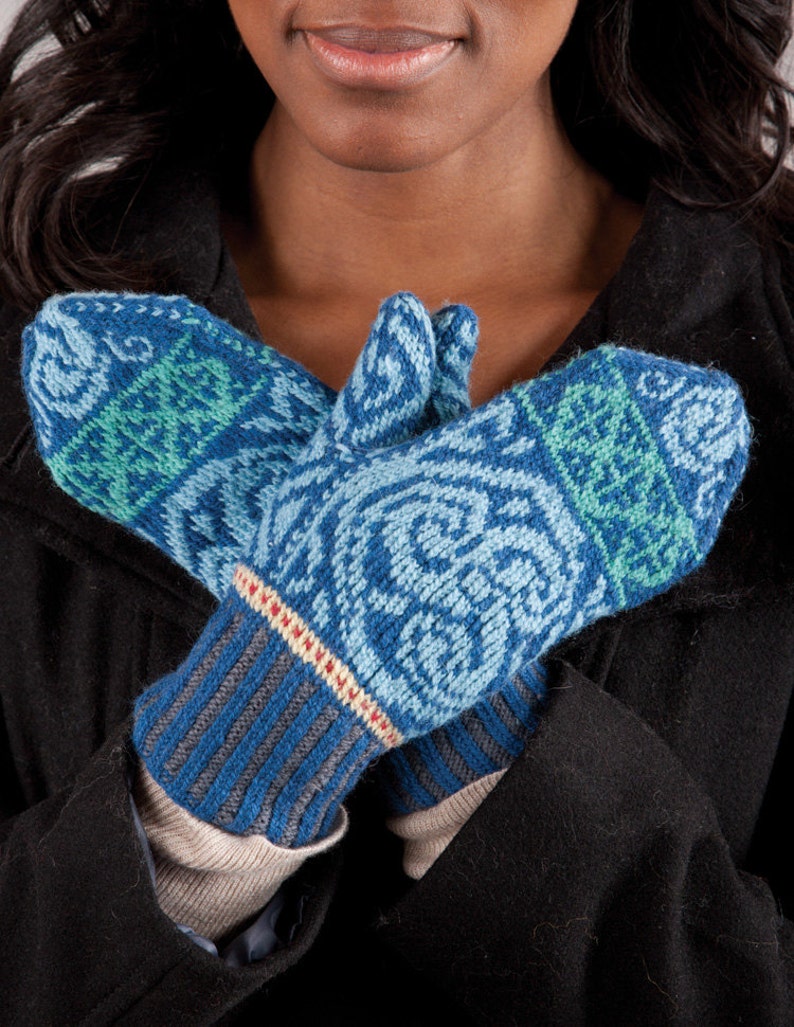 May include: A pair of blue and green knitted mittens with a white and red stripe detail. The mittens are held together in front of a person wearing a black coat.