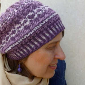 May include: A close-up of a woman wearing a purple and white knitted hat with a geometric pattern. The hat is pulled down over her forehead and she is looking down. She is wearing a light brown scarf and has a purple earring.