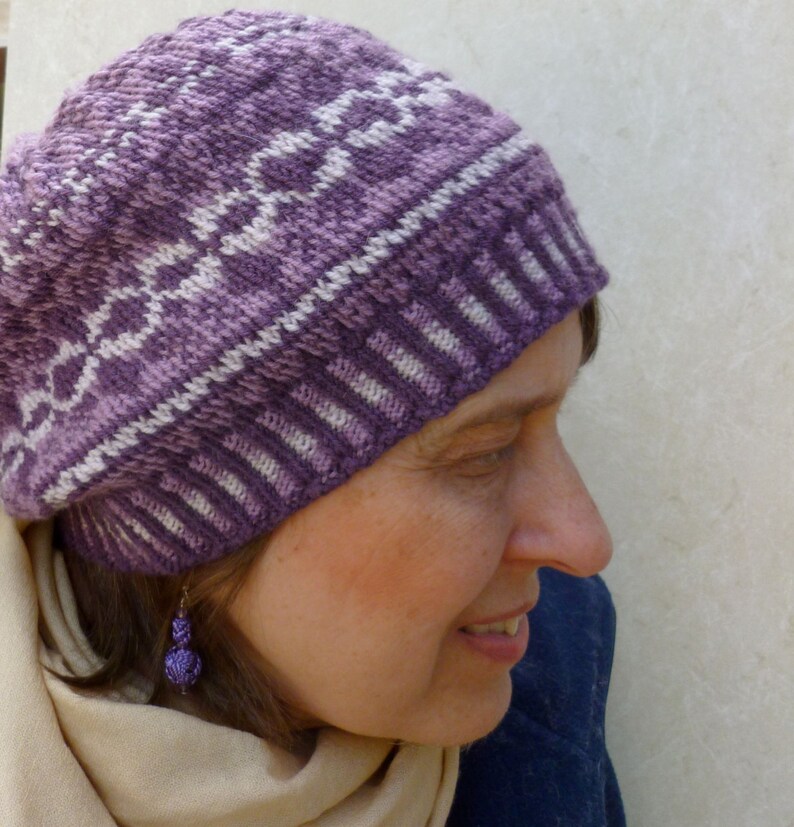 May include: A close-up of a woman wearing a purple and white knitted hat with a geometric pattern. The hat is pulled down over her forehead and she is looking down. She is wearing a light brown scarf and has a purple earring.