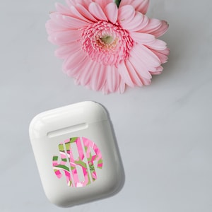 May include: White Airpod case with a pink and green floral monogram sticker.