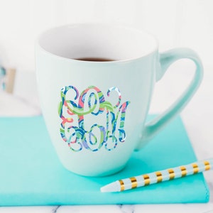 May include: A light blue ceramic mug with a white interior, featuring a colorful, swirling, multi-patterned monogram design on the front. The mug is positioned on a bright turquoise notebook next to a light blue and gold striped pen.