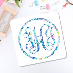 May include: A square white coaster featuring a circular design with a swirling, multi-colored pattern in shades of blue, pink, green, and purple. Inside the circle is an ornate, interlocking monogram featuring the letters H and G in the same pattern.