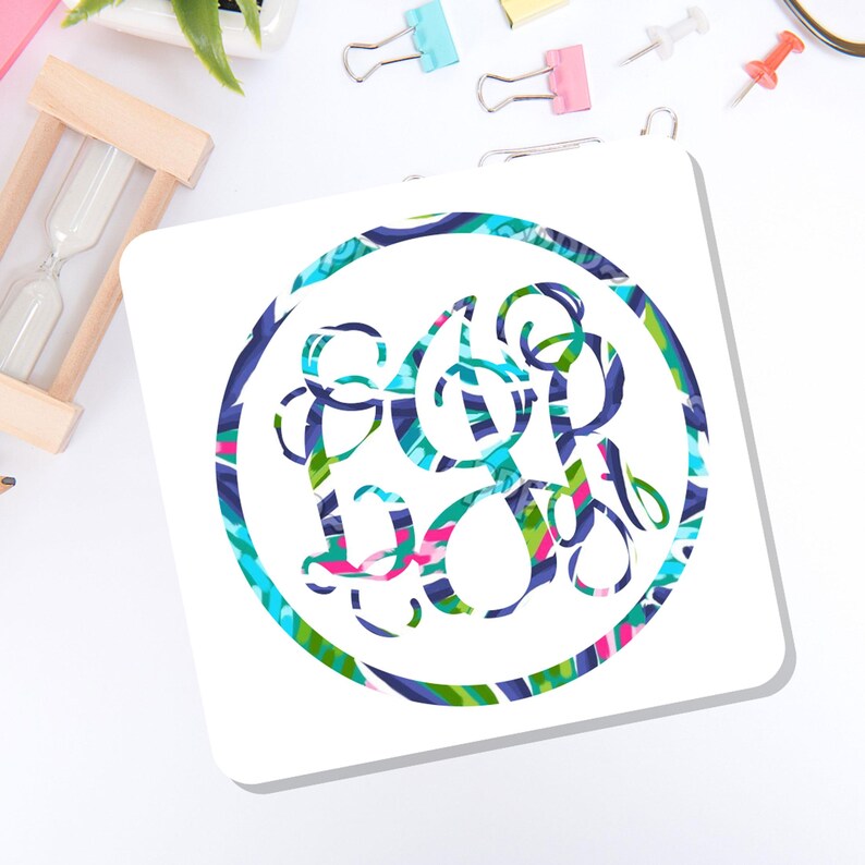 May include: A square white coaster featuring a circular design filled with a vibrant, multi-colored abstract pattern of blue, green, pink, and white swirls. The central graphic contains an intricate, stylized monogram with overlapping letters.