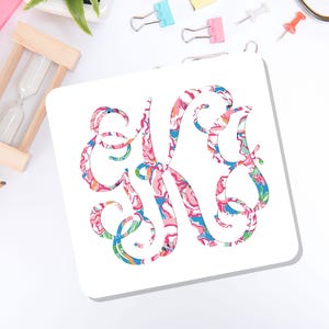 May include: A square white coaster featuring a large, ornate, swirling monogram letter K. The letter is filled with a vibrant, multi-colored floral and abstract pattern in shades of pink, blue, green, and orange, set against a plain white background.