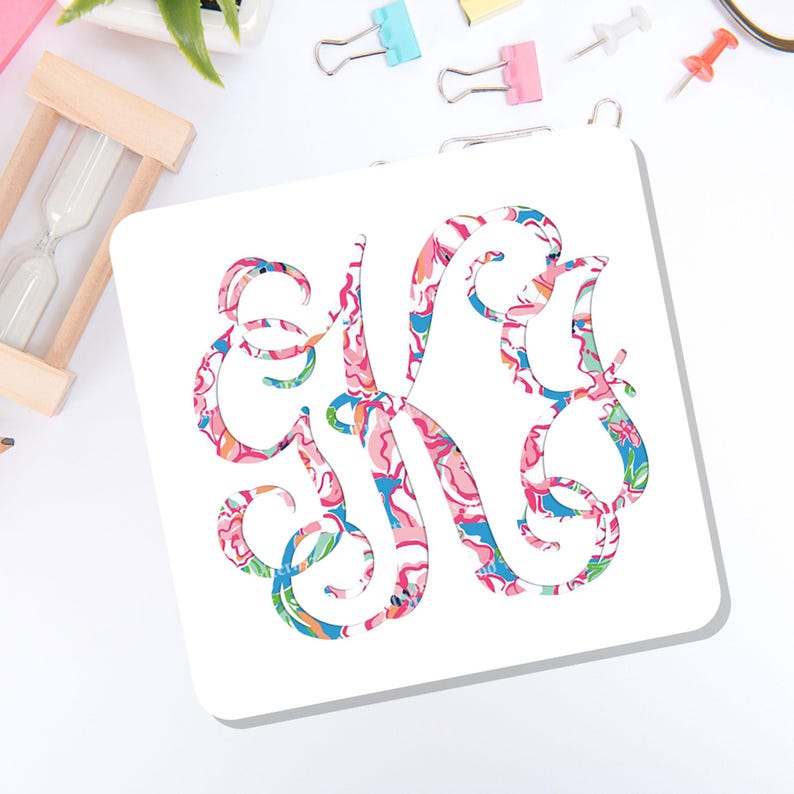 May include: A square white coaster featuring a large, ornate, swirling monogram letter K. The letter is filled with a vibrant, multi-colored floral and abstract pattern in shades of pink, blue, green, and orange, set against a plain white background.