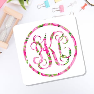 May include: A square coaster featuring a circular design with a decorative, swirling monogram letter K. The pattern consists of a vibrant, abstract mix of bright pink, lime green, and white colors, creating a bold and colorful visual effect.