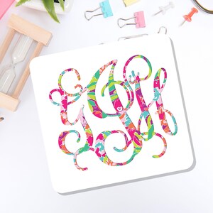 May include: A square white coaster featuring a large, ornate, interlocking monogram in the center. The letters are filled with a vibrant, multi-colored floral and abstract pattern in shades of hot pink, bright green, turquoise, and orange.