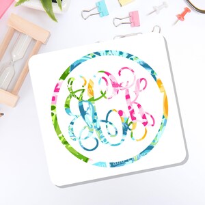 May include: A square white coaster featuring a circular design in the center. The design consists of a colorful, multi-patterned monogram enclosed within a circular border that matches the vibrant, abstract floral and geometric print of the letters.