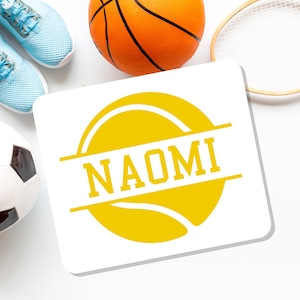 May include: A white square coaster with the name "NAOMI" in yellow, surrounded by a yellow circle and horizontal lines. The coaster is next to a basketball, a soccer ball, a tennis racket, and a pair of light blue sneakers.