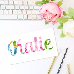 May include: A white card with the name "Katie" in a colorful floral pattern. The card is surrounded by a keyboard, pink and white flowers, and gold and black pens. The floral design includes pink, green, blue, and yellow.