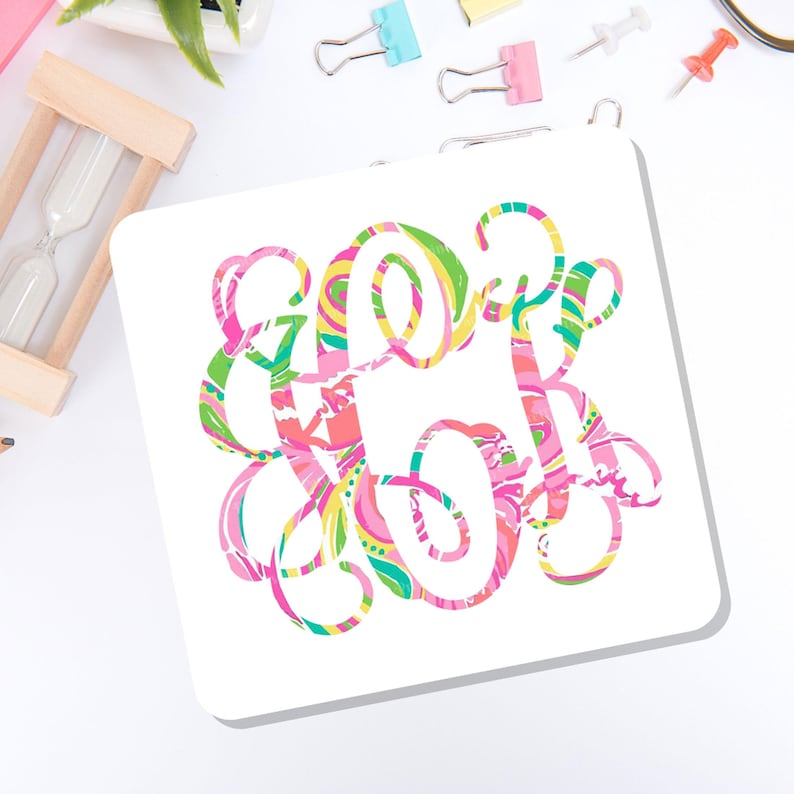 May include: A square white coaster featuring a decorative, swirling monogram design in vibrant shades of pink, yellow, lime green, and turquoise. The intricate, interconnected letters create a colorful, abstract pattern centered on the surface.