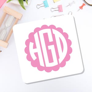 May include: A square white coaster featuring a pink scalloped circular border surrounding a white monogram with the letters HGD in a bold, block-style font. The design is centered on the coaster, which has rounded corners and a clean, flat surface.
