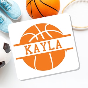 May include: A white square coaster with an orange basketball design and the name "KAYLA" in orange block letters. Other sports equipment, including a basketball, tennis racket, soccer ball, and blue sneakers, are visible in the background.