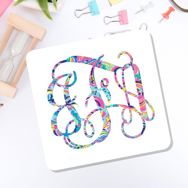 May include: A square white coaster featuring a large, ornate, interlocking monogram design in the center. The monogram is filled with a vibrant, multi-colored abstract pattern consisting of swirls and shapes in shades of pink, blue, yellow, and green.