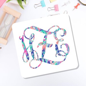 May include: A square white coaster featuring a large, ornate, interlocking monogram in a vibrant, multi-colored abstract pattern. The design incorporates swirling shades of pink, blue, teal, and purple in a fluid, painterly style across the letters.