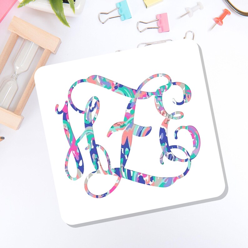 May include: A square white coaster featuring a large, ornate, interlocking monogram in a vibrant, multi-colored abstract pattern. The design incorporates swirling shades of pink, blue, teal, and purple in a fluid, painterly style across the letters.
