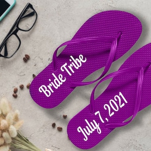 May include: A pair of purple flip flops with white text. One flip flop says "Bride Tribe" and the other says "July 7, 2021".