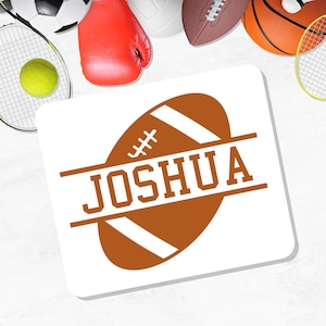 May include: A white square coaster with a brown football design and the name "JOSHUA" in bold letters. Various sports equipment, including a tennis racket, ball, and boxing glove, are visible in the background.