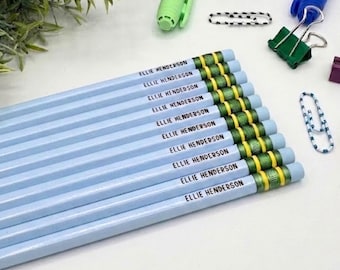 May include: A set of light blue pencils, each featuring the name ELLIE HENDERSON printed in black capital letters. The pencils have green and gold metallic ferrules at the ends, arranged in a parallel row alongside various office supplies.