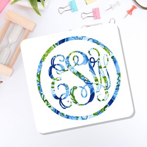 May include: A square white coaster featuring a circular design in the center. The circular graphic contains an intricate, swirling monogram with the letters S and W, decorated in a vibrant pattern of blue, light blue, and green floral motifs.