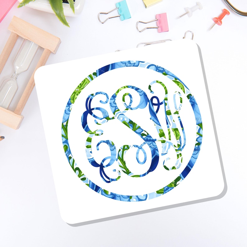 May include: A square white coaster featuring a circular design in the center. The circular graphic contains an intricate, swirling monogram with the letters S and W, decorated in a vibrant pattern of blue, light blue, and green floral motifs.