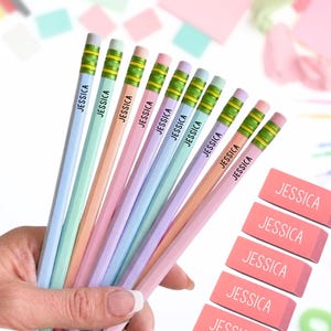May include: A set of pastel-colored pencils and rectangular erasers, each personalized with the name JESSICA printed in black or white. The pencils feature green metal ferrules and pink erasers, arranged in a fan shape alongside stacked erasers.