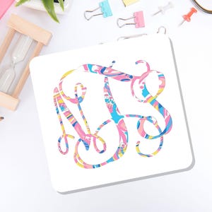 May include: A square white coaster featuring a decorative, swirling monogram design in the center. The letters are filled with a vibrant, multi-colored pattern consisting of pink, blue, yellow, and white abstract shapes and wavy lines.