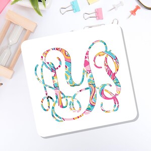 May include: A square white coaster featuring a large, ornate, interlocking monogram letter design. The monogram is filled with a vibrant, multi-colored paisley-inspired pattern in shades of bright pink, turquoise blue, yellow, and orange.