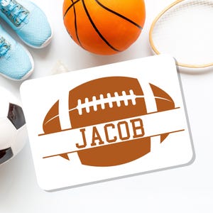May include: A white rectangular sign featuring a brown football design with the name "JACOB" in white. Surrounding the sign are a basketball, soccer ball, tennis racket, and light blue sneakers.