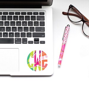 May include: A silver laptop computer with a colorful monogram sticker on the lid. A pink pen and brown-framed glasses are on a white surface next to the laptop.