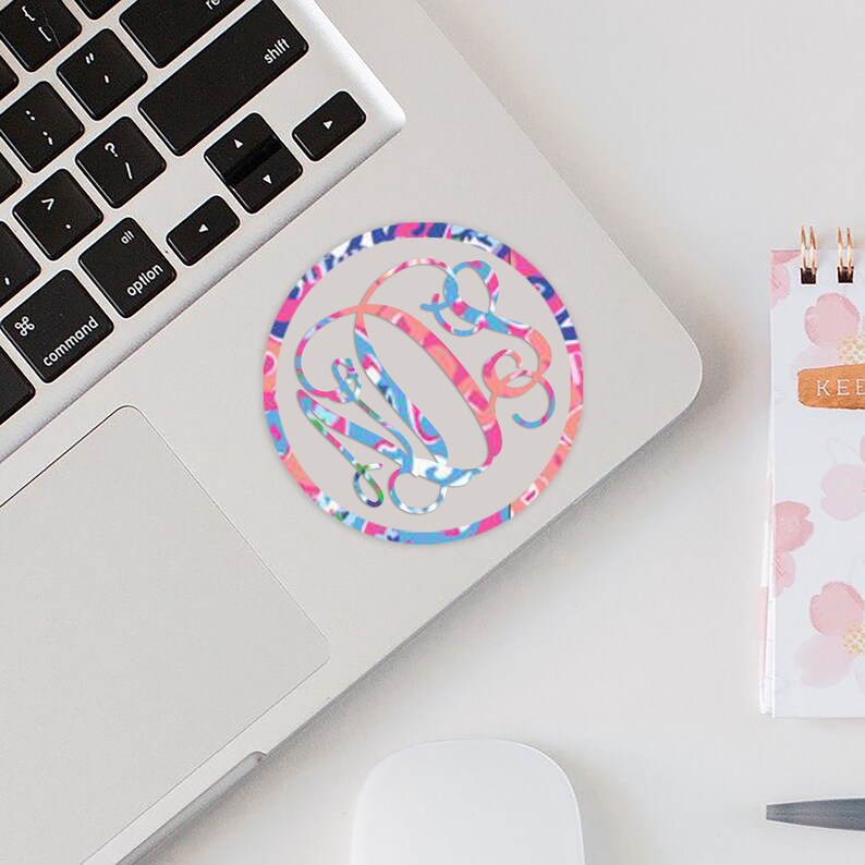 May include: A pink, blue, and white patterned circle sticker with a white monogram in the center. The sticker is on a silver laptop. Pattern 7