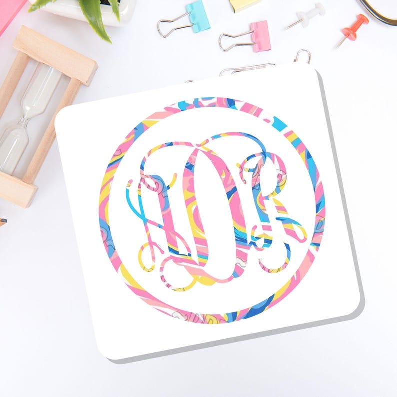 May include: A square white coaster featuring a circular design with a colorful marbled pattern in shades of pink, yellow, and blue. The center of the circle displays an ornate, intertwined monogram with the letters D and R in the same marbled style.
