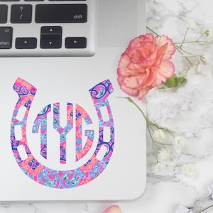May include: A pink and blue horseshoe-shaped sticker with a monogram in the center. The sticker is on a white laptop.