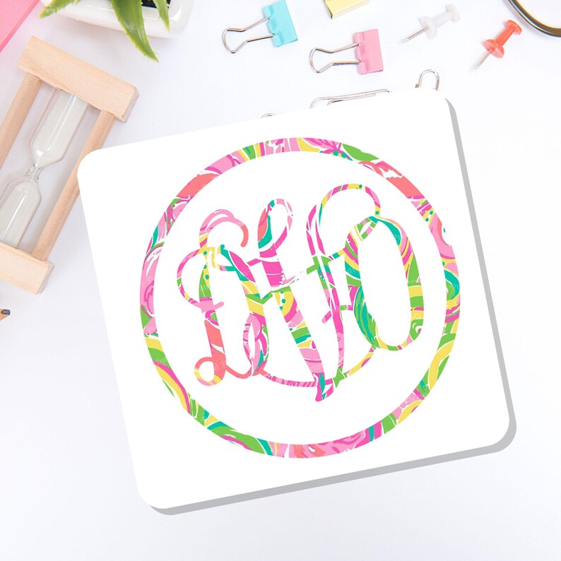 May include: A square white coaster featuring a circular design with a colorful, swirling pattern in shades of pink, green, and yellow. Inside the circle is a stylized, interlocking monogram of the letters L, F, and O in the same vibrant, multi-colored print.