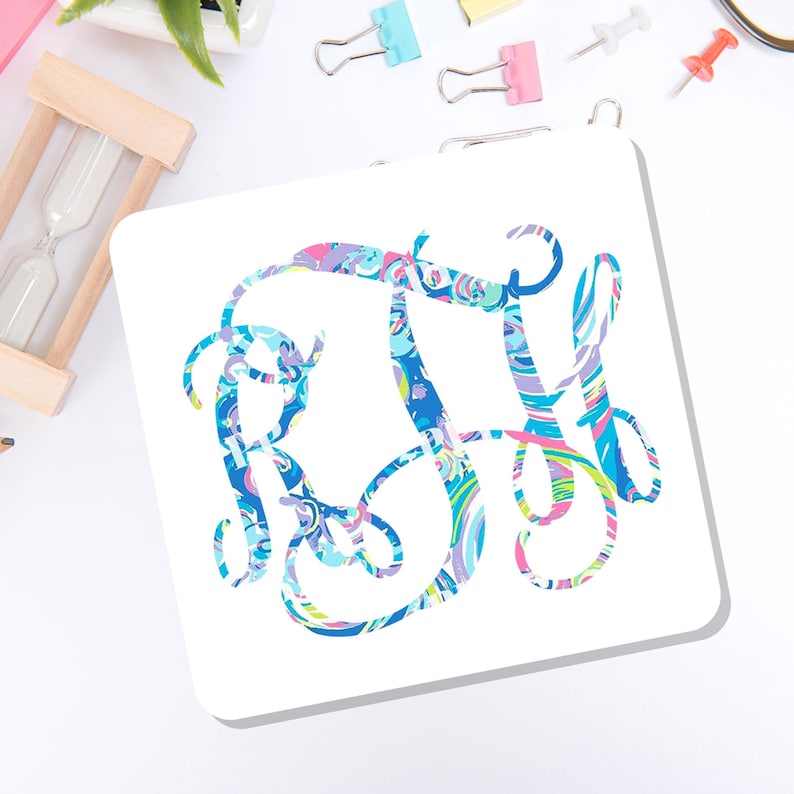 May include: A square white coaster featuring a decorative, swirling monogram in shades of blue, pink, purple, and green. The letters are intertwined in a cursive, ornate script style, creating a colorful, abstract pattern across the center.