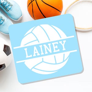 May include: A light blue square coaster with a white volleyball design and the name "LAINEY" in white text. The coaster is surrounded by sports equipment, including a basketball, tennis racket, soccer ball, and light blue sneakers.