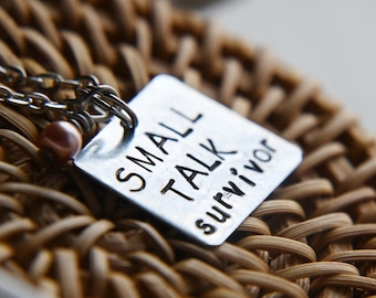 Könnte beinhalten: Halskette mit quadratischem Anhänger aus Silber, auf dem in schwarzer Schrift "SMALL TALK survivor" steht. Die Kette hat eine kleine, runde, rosa Perle. Der Anhänger liegt auf einer gewebten, braunen Oberfläche.