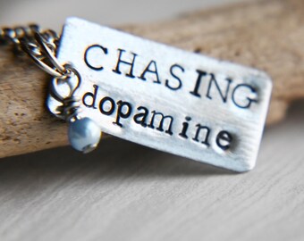 Könnte beinhalten: Silberne Halskette mit einem Anhänger, auf dem "CHASING dopamine" steht. Der Anhänger ist an einer silbernen Kette mit einer kleinen blauen Perle befestigt.