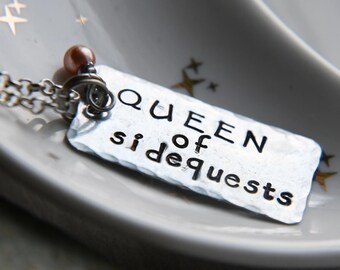 Könnte beinhalten: Eine silberne Halskette mit einem Anhänger, auf dem "QUEEN of sidequests" steht.